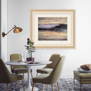 May include: A framed photograph of a wave crashing on a beach at sunset. The photo is in a light wood frame with a white mat. The photo is hanging on a white wall above a dining table with four chairs.