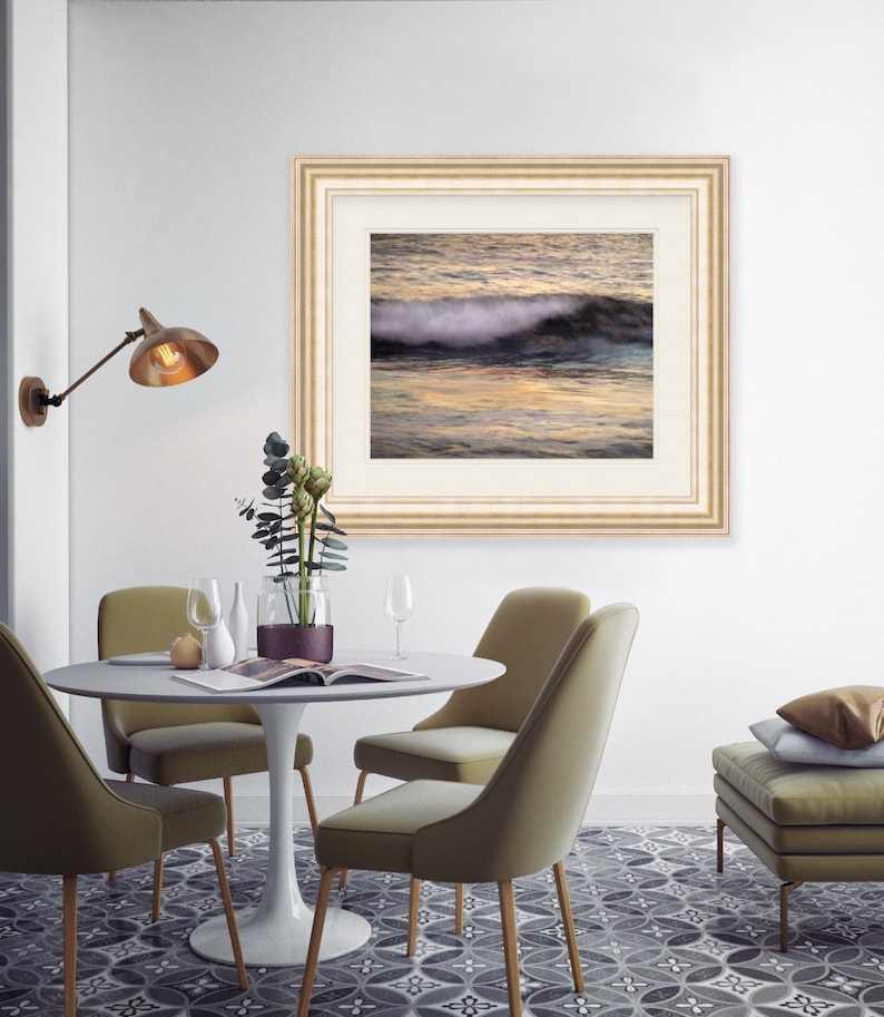 May include: A framed photograph of a wave crashing on a beach at sunset. The photo is in a light wood frame with a white mat. The photo is hanging on a white wall above a dining table with four chairs.