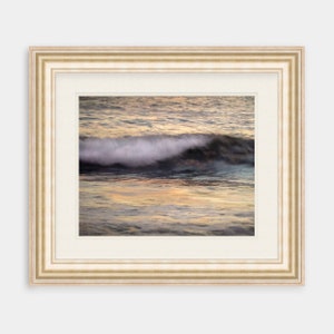 May include: A framed photograph of a wave crashing in the ocean at sunset. The water is a mix of blue, green, and gold, and the wave is white and foamy. The frame is a simple, off-white color.