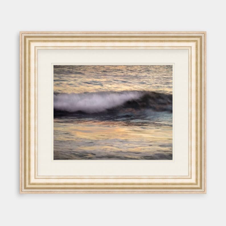 May include: A framed photograph of a wave crashing in the ocean at sunset. The water is a mix of blue, green, and gold, and the wave is white and foamy. The frame is a simple, off-white color.