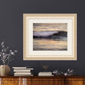 May include: A framed photograph of a wave crashing in the ocean at sunset. The water is a mix of blue, green, and orange hues. The frame is a light beige color with a white mat.