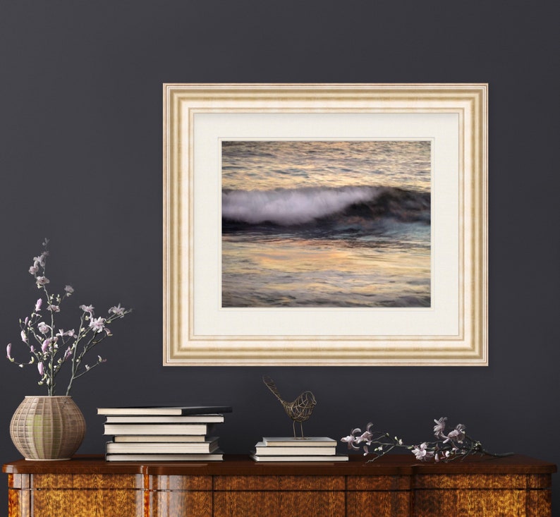 May include: A framed photograph of a wave crashing in the ocean at sunset. The water is a mix of blue, green, and orange hues. The frame is a light beige color with a white mat.