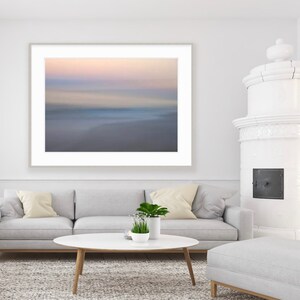 May include: A white couch with two beige pillows sits in front of a framed abstract photograph of a blurred beach scene. A white oval coffee table with a small potted plant sits in front of the couch. A white ottoman sits in the foreground.