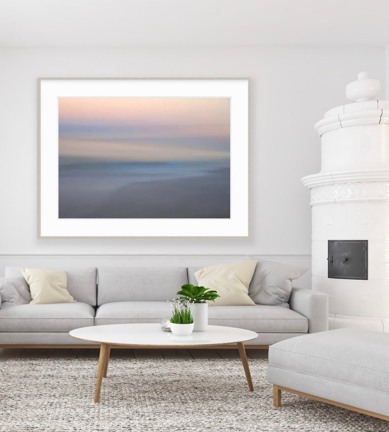 May include: A white couch with two beige pillows sits in front of a framed abstract photograph of a blurred beach scene. A white oval coffee table with a small potted plant sits in front of the couch. A white ottoman sits in the foreground.