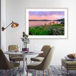 May include: A framed photograph of a lake with a pink and purple sunset sky. The lake is surrounded by green trees and bushes. There are yellow and purple flowers in the foreground.
