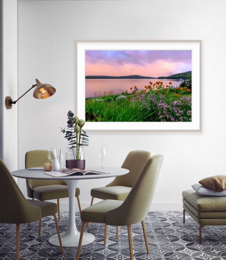 May include: A framed photograph of a lake with a pink and purple sunset sky. The lake is surrounded by green trees and bushes. There are yellow and purple flowers in the foreground.