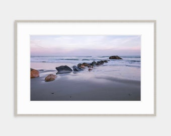 May include: A line of rocks extends into the ocean at low tide. The rocks are dark gray and wet, and the sand is light gray and wet. The sky is a pale blue with a hint of pink in the distance.