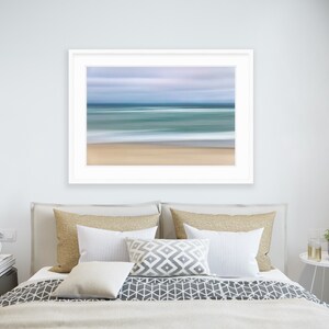 May include: A framed abstract photograph of a beach scene with blurred lines of blue, green, and white representing the ocean and sky. The sand is a light brown color.