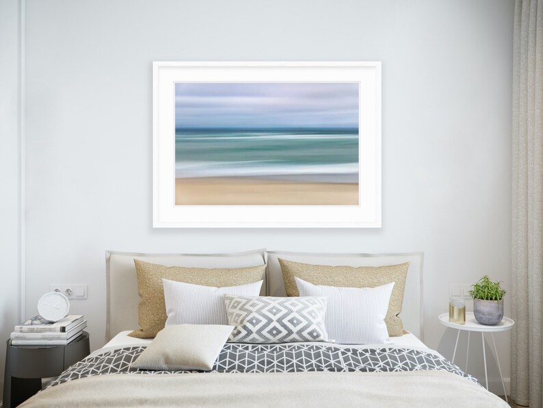 May include: A framed abstract photograph of a beach scene with blurred lines of blue, green, and white representing the ocean and sky. The sand is a light brown color.