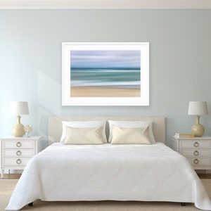 May include: A white bed with a white comforter and two beige pillows in a bedroom with light blue walls. A framed abstract seascape painting hangs above the bed. Two white nightstands with drawers are on either side of the bed, each with a lamp with a white shade and a beige base.