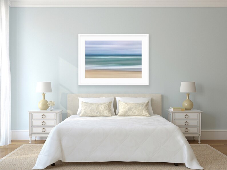 May include: A white bed with a white comforter and two beige pillows in a bedroom with light blue walls. A framed abstract seascape painting hangs above the bed. Two white nightstands with drawers are on either side of the bed, each with a lamp with a white shade and a beige base.