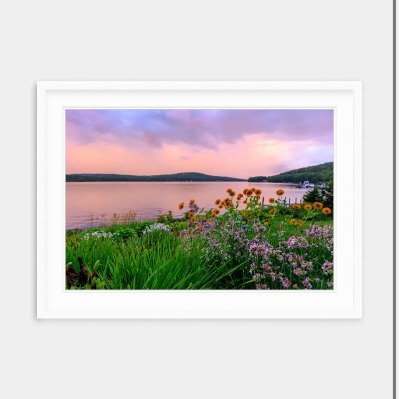 May include: A scenic landscape photograph of a lake with a mountain range in the background. The lake is calm and still, and the sky is a soft pink and purple. There are flowers in the foreground, and the image is framed by a white mat.