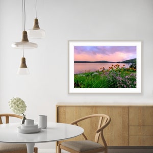 May include: A framed photograph of a lake with a pink and purple sunset sky. The lake is surrounded by green trees and bushes with pink and yellow flowers.