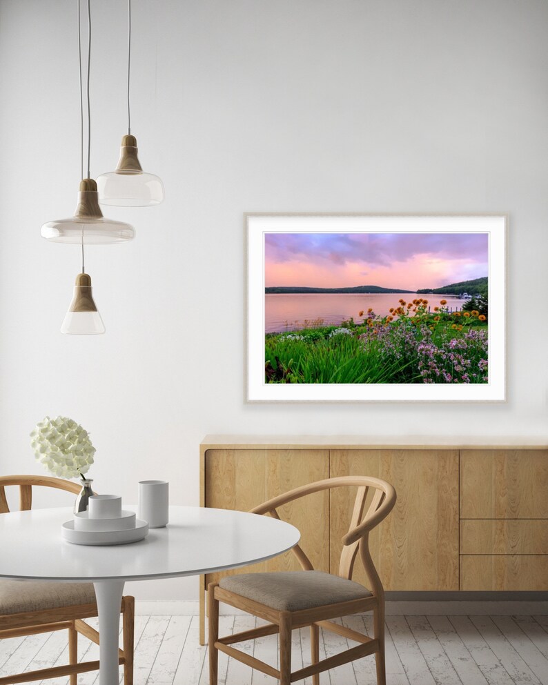 May include: A framed photograph of a lake with a pink and purple sunset sky. The lake is surrounded by green trees and bushes with pink and yellow flowers.