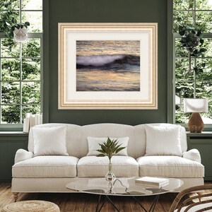 May include: A framed photograph of a wave crashing on a beach at sunset. The water is a mix of blue, green, and orange, and the sand is a light brown. The frame is a light wood color.