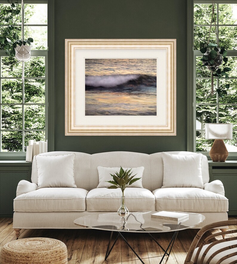 May include: A framed photograph of a wave crashing on a beach at sunset. The water is a mix of blue, green, and orange, and the sand is a light brown. The frame is a light wood color.