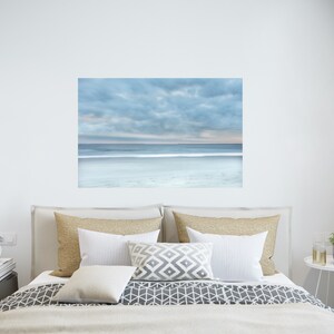 May include: A bedroom interior design featuring a large framed photograph of a beach scene with a cloudy sky and a calm ocean. The photograph is hanging above a bed with a white duvet cover and a grey and white patterned bedspread. There are two beige pillows and a white pillow on the bed. A small white bedside table with a clock and books is next to the bed. A small white bedside table with a plant is on the right side of the bed.