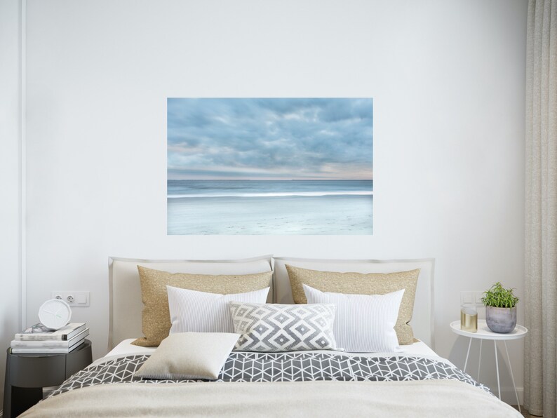 May include: A bedroom interior design featuring a large framed photograph of a beach scene with a cloudy sky and a calm ocean. The photograph is hanging above a bed with a white duvet cover and a grey and white patterned bedspread. There are two beige pillows and a white pillow on the bed. A small white bedside table with a clock and books is next to the bed. A small white bedside table with a plant is on the right side of the bed.