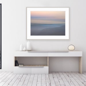 May include: A white console table with a framed photograph of a blurred seascape with pink and blue hues. The table has two shelves and is decorated with three white ceramic vases. A black floor lamp with a white lampshade is on the right side of the image.