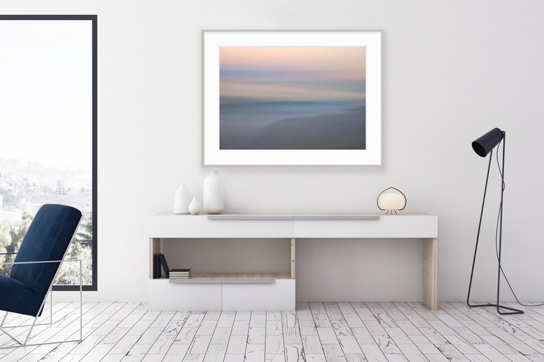 May include: A white console table with a framed photograph of a blurred seascape with pink and blue hues. The table has two shelves and is decorated with three white ceramic vases. A black floor lamp with a white lampshade is on the right side of the image.