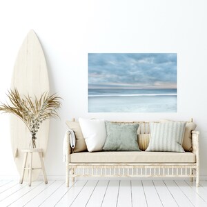 May include: A light blue and white beach scene photograph with a cloudy sky and a white sandy beach. The photograph is hanging on a white wall above a light brown couch with white and green cushions.