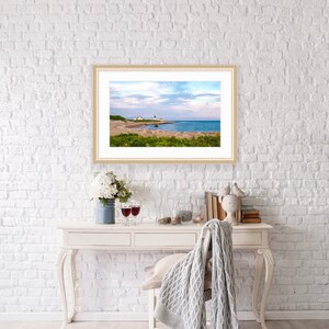 May include: A framed photograph of a lighthouse on a rocky coastline with a blue sky and white clouds. The lighthouse is white with a red roof and is located on a small island. The water is blue and the sky is a light blue with white clouds.