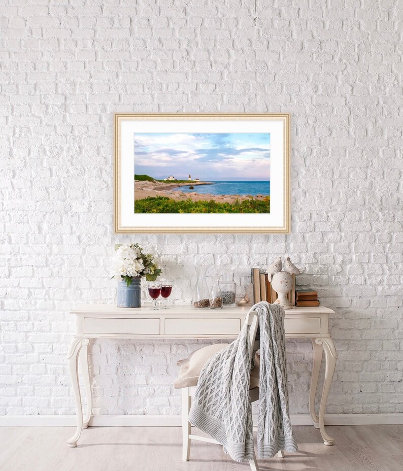 May include: A framed photograph of a lighthouse on a rocky coastline with a blue sky and white clouds. The lighthouse is white with a red roof and is located on a small island. The water is blue and the sky is a light blue with white clouds.