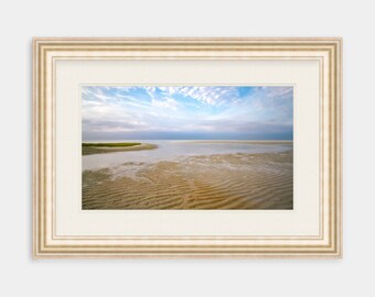 May include: A framed photograph of a beach scene with a blue sky, white clouds, and a sandy beach with ripples in the sand. The water is calm and the horizon is in the distance.