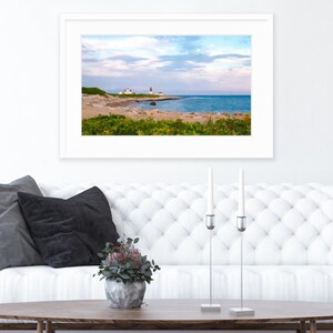 May include: A framed photograph of a lighthouse on a rocky coastline with a blue ocean and cloudy sky. The photograph is hanging on a white wall above a white tufted sofa with black pillows. A vase with green plants sits on a wooden coffee table in front of the sofa.