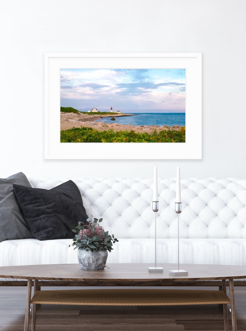 May include: A framed photograph of a lighthouse on a rocky coastline with a blue ocean and cloudy sky. The photograph is hanging on a white wall above a white tufted sofa with black pillows. A vase with green plants sits on a wooden coffee table in front of the sofa.
