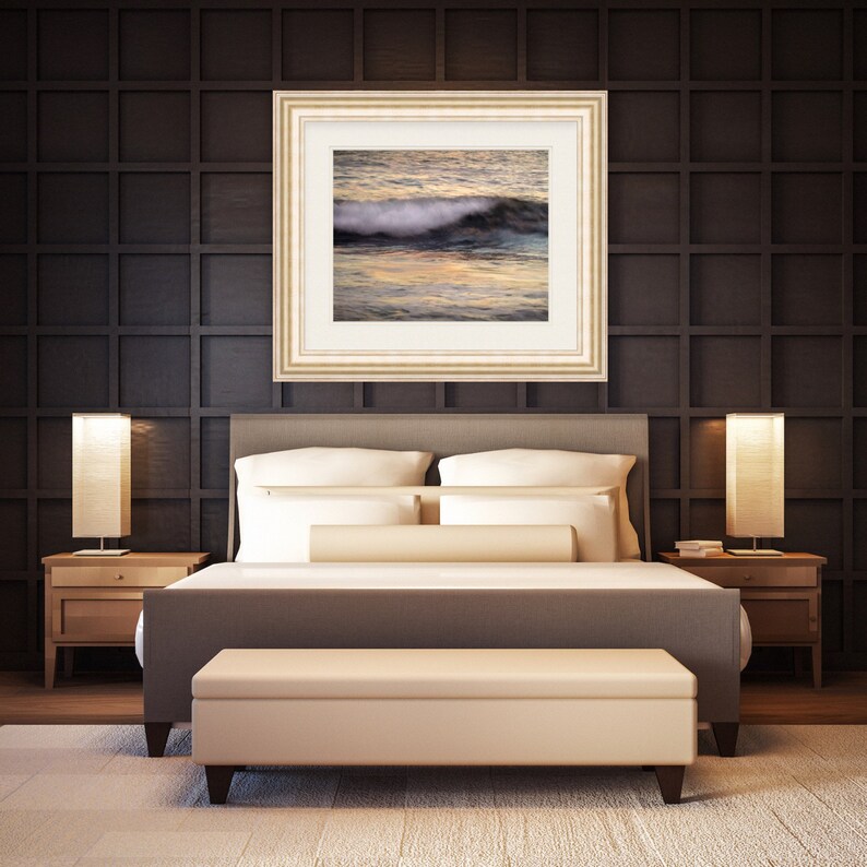 May include: A bedroom with a king-sized bed with white bedding and a framed photograph of a wave crashing on a beach above the headboard. There are two nightstands with lamps on either side of the bed.