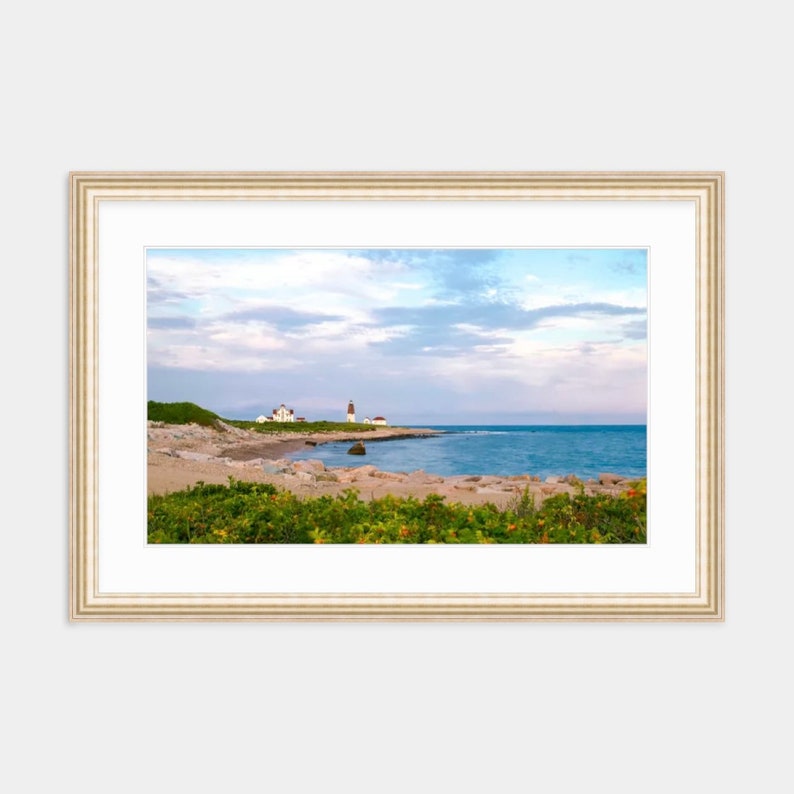 May include: A photograph of a lighthouse on a rocky coastline with a sandy beach. The sky is a light blue with white clouds. The water is a deep blue.