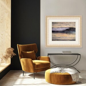 May include: A framed photograph of a wave crashing on a beach at sunset. The photo is in a white frame with a gold trim and is hanging on a white wall. A mustard yellow armchair and ottoman are in the foreground.