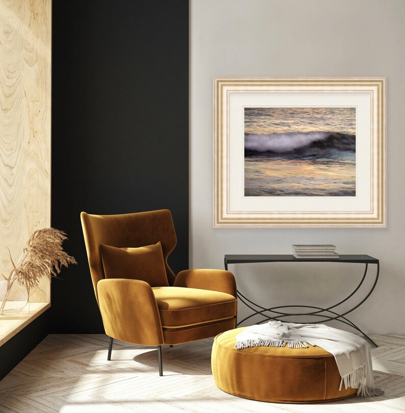 May include: A framed photograph of a wave crashing on a beach at sunset. The photo is in a white frame with a gold trim and is hanging on a white wall. A mustard yellow armchair and ottoman are in the foreground.