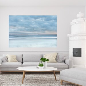 May include: A photograph of a beach scene with a cloudy sky and a calm ocean. The photo is printed on canvas and framed in a white frame. The photo is hanging on a white wall above a grey sofa and a white coffee table.