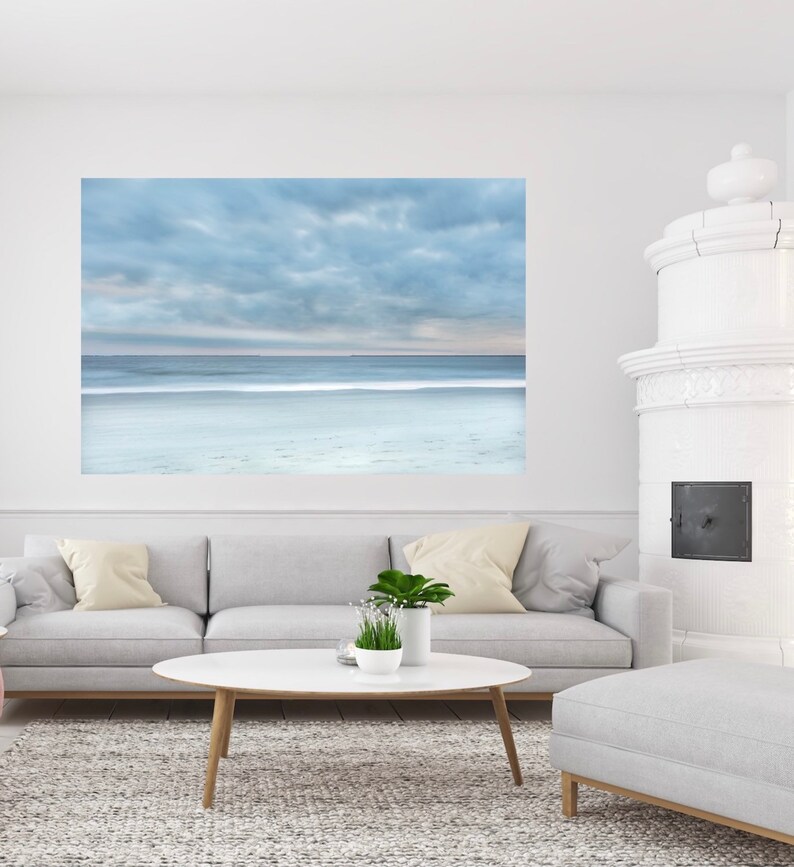May include: A photograph of a beach scene with a cloudy sky and a calm ocean. The photo is printed on canvas and framed in a white frame. The photo is hanging on a white wall above a grey sofa and a white coffee table.