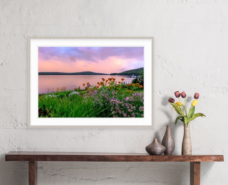 May include: A framed photograph of a lake with a pink and purple sunset sky. The lake is surrounded by green trees and bushes with yellow and purple flowers in the foreground.