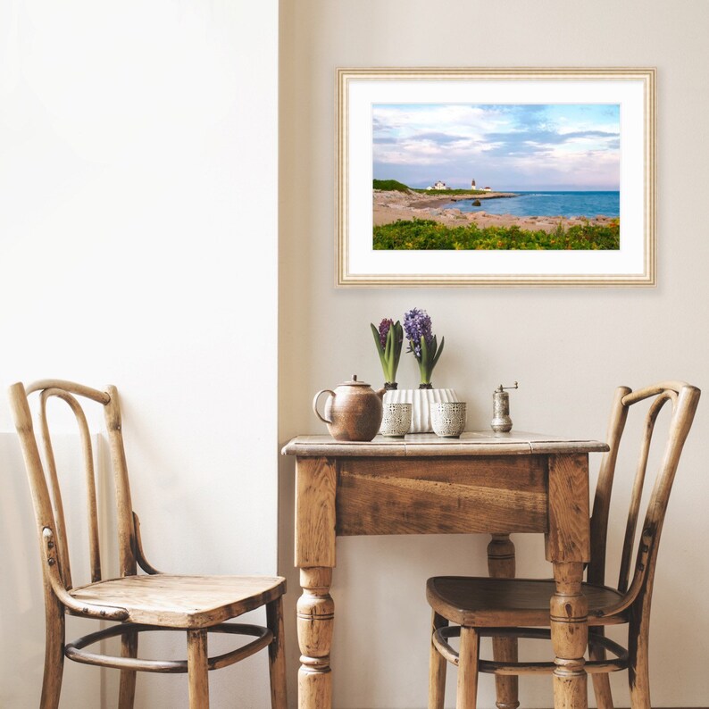 May include: A framed photograph of a coastal scene with a lighthouse in the distance. The photo is hanging on a white wall above a wooden table with two chairs. There are two vases with purple flowers on the table.