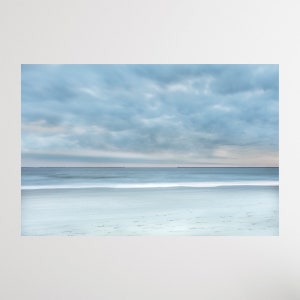 May include: A panoramic view of a sandy beach with a calm, blue ocean and a cloudy sky. The water is a light blue and the sand is a light beige. The sky is a light blue with white clouds.