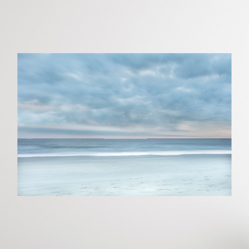 May include: A panoramic view of a sandy beach with a calm, blue ocean and a cloudy sky. The water is a light blue and the sand is a light beige. The sky is a light blue with white clouds.