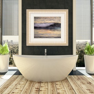 May include: A white bathtub with a gold faucet in a modern bathroom with wood floors. A framed picture of a wave crashing on a beach hangs on the wall behind the tub. Two potted plants are on either side of the tub.