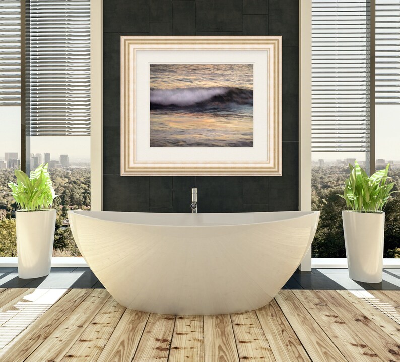 May include: A white bathtub with a gold faucet in a modern bathroom with wood floors. A framed picture of a wave crashing on a beach hangs on the wall behind the tub. Two potted plants are on either side of the tub.