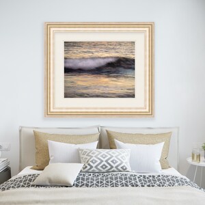 May include: A framed photograph of a wave crashing on a beach at sunset. The water is a deep blue and the sky is a soft orange and pink. The wave is white and foamy.