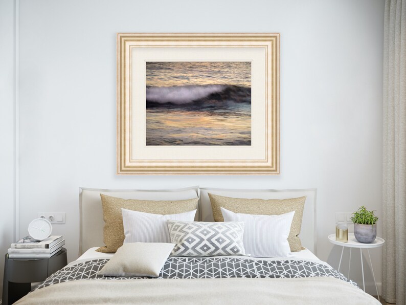 May include: A framed photograph of a wave crashing on a beach at sunset. The water is a deep blue and the sky is a soft orange and pink. The wave is white and foamy.