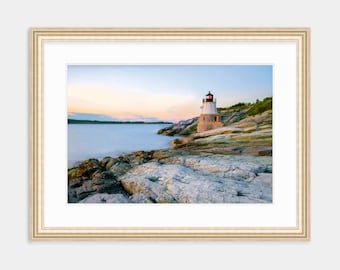 May include: A white lighthouse with a black roof stands on a rocky shore at sunset. The water is calm and the sky is a soft blue.
