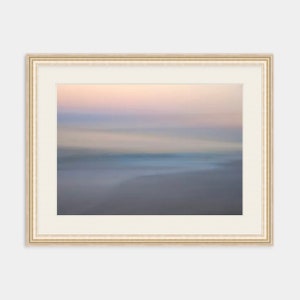 May include: A framed abstract painting of a blurred seascape with soft pink, blue, and gray hues. The frame is a light beige color with a subtle raised edge.