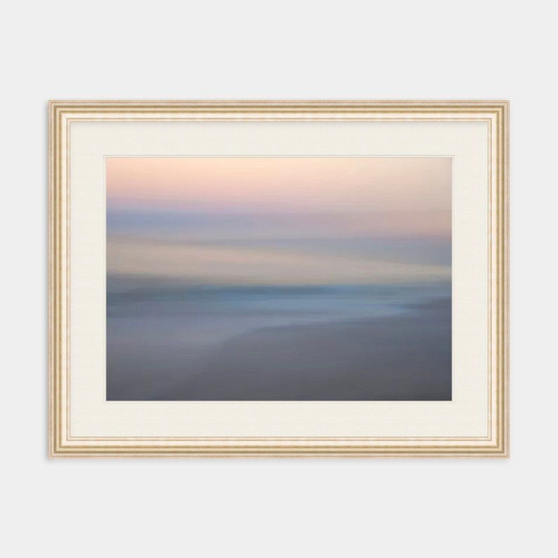 May include: A framed abstract painting of a blurred seascape with soft pink, blue, and gray hues. The frame is a light beige color with a subtle raised edge.