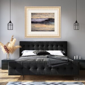 May include: A framed photograph of a wave crashing on a beach at sunset. The water is a deep blue and the sky is a soft orange and pink. The photo is in a light wood frame.