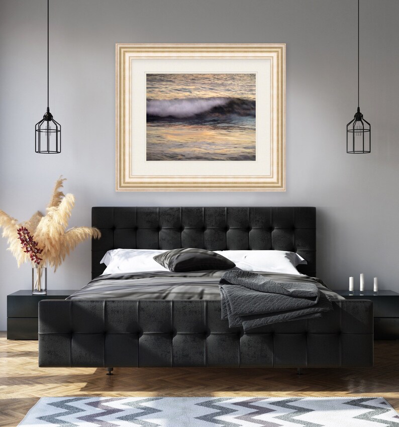 May include: A framed photograph of a wave crashing on a beach at sunset. The water is a deep blue and the sky is a soft orange and pink. The photo is in a light wood frame.