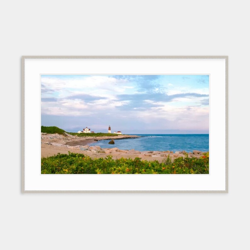 May include: A white lighthouse stands on a rocky shore with a sandy beach in the foreground. The sky is a soft blue with fluffy white clouds. The ocean is calm and blue.